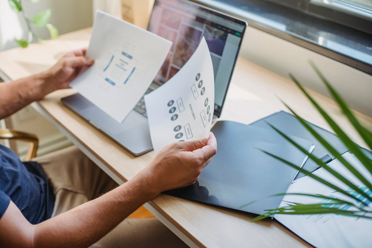 Home Crop anonymous male designer in casual wear sitting at table with opened laptop and folders and reading papers